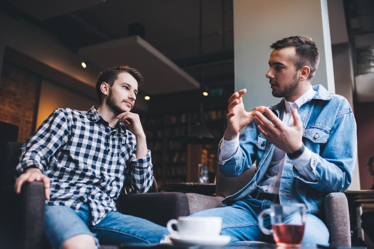 From below young handsome modern men in casual outfit sitting on chairs at table with tea and coffee discussing together life matters in modern cafe.