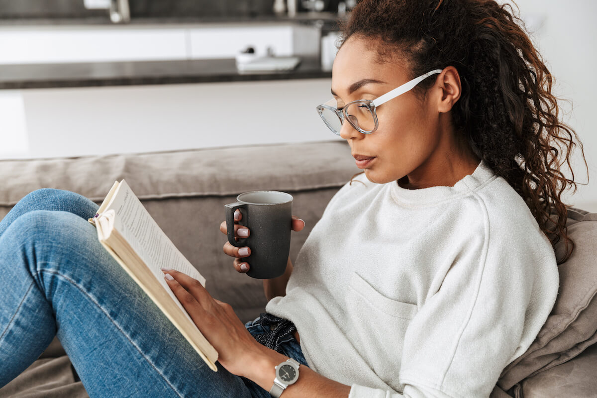 African american girl reading book and drinking tea while sitting on sofa in bright flat.