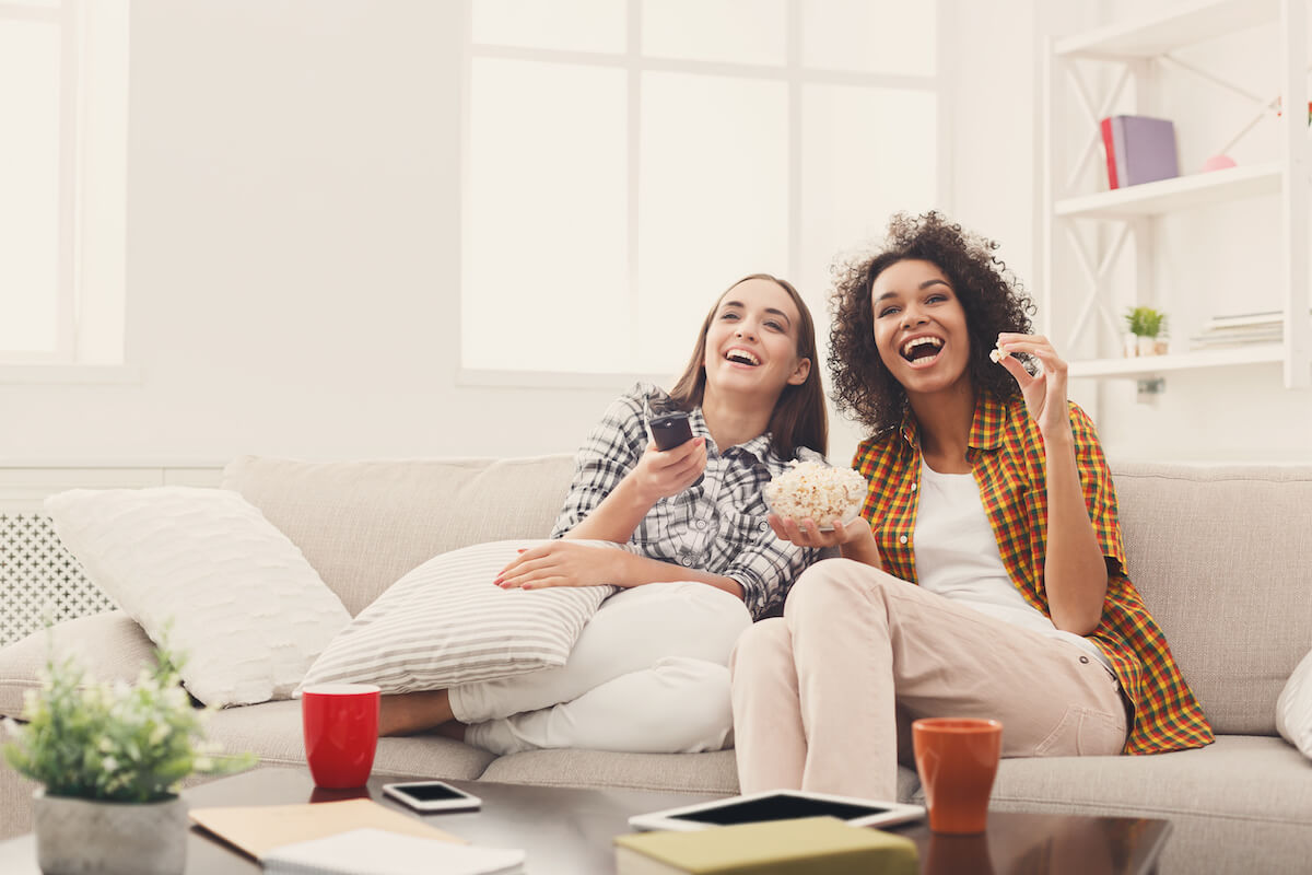 Smiling female friends relaxing and watching TV at home, eating popcorn