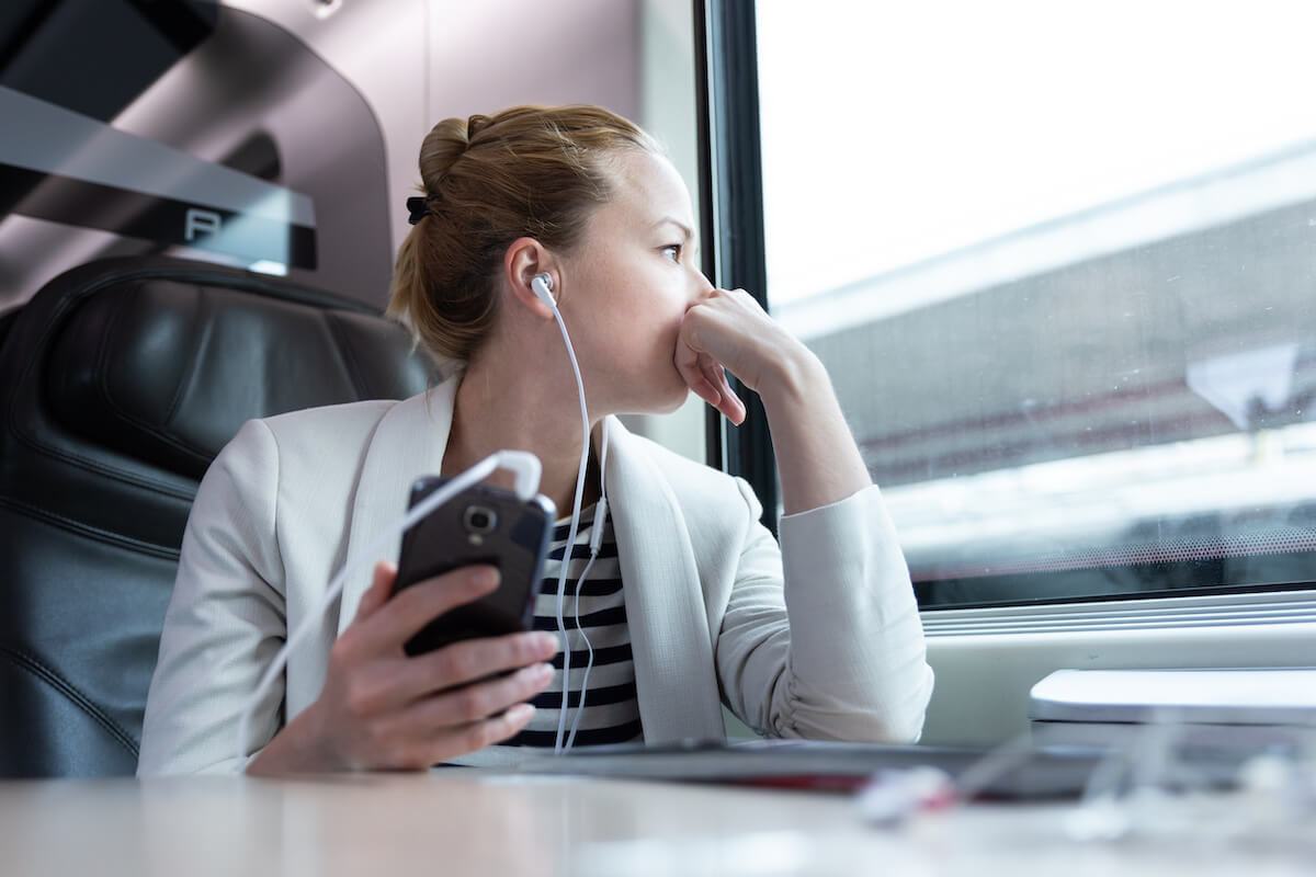 Thoughtful businesswoman looking trough the window, listening to podcast on cellphone using headphone set while traveling by train in business class seat.