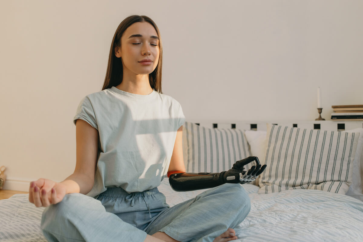 Pretty cyber lady with 3d-printed bionic hand prosthesis doing yoga sitting in lotus posture, fingers in mudra sign, having eyes closed, meditating on bed in morning