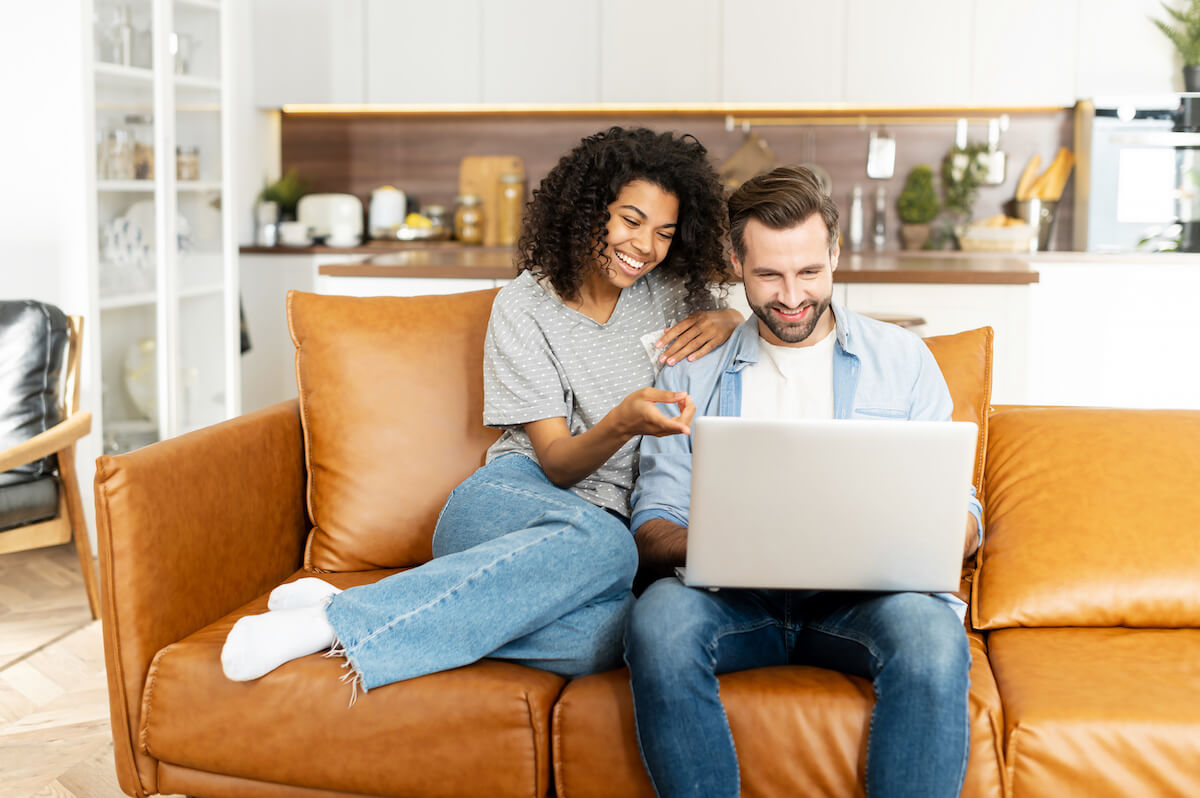 Cheerful diverse couple in love spends leisure time online with a laptop at home.