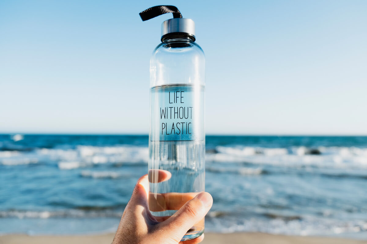 Closeup of a caucasian man holding a glass reusable water bottle with the text life without plastic written in it, on the beach, with the ocean in the background.
