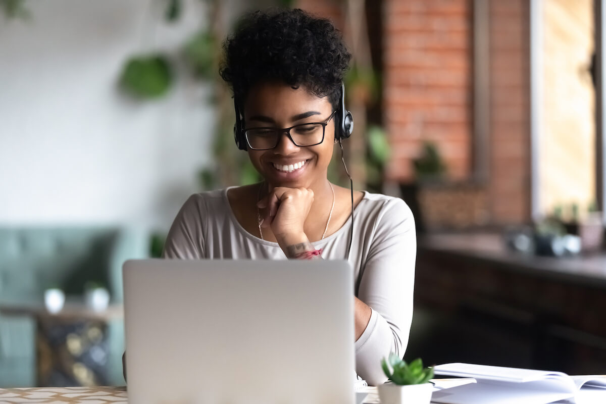 Black woman smart student girl sitting at table in university cafe alone wearing glasses looking at computer screen using headphones listening online lecture improve language skills having good mood.