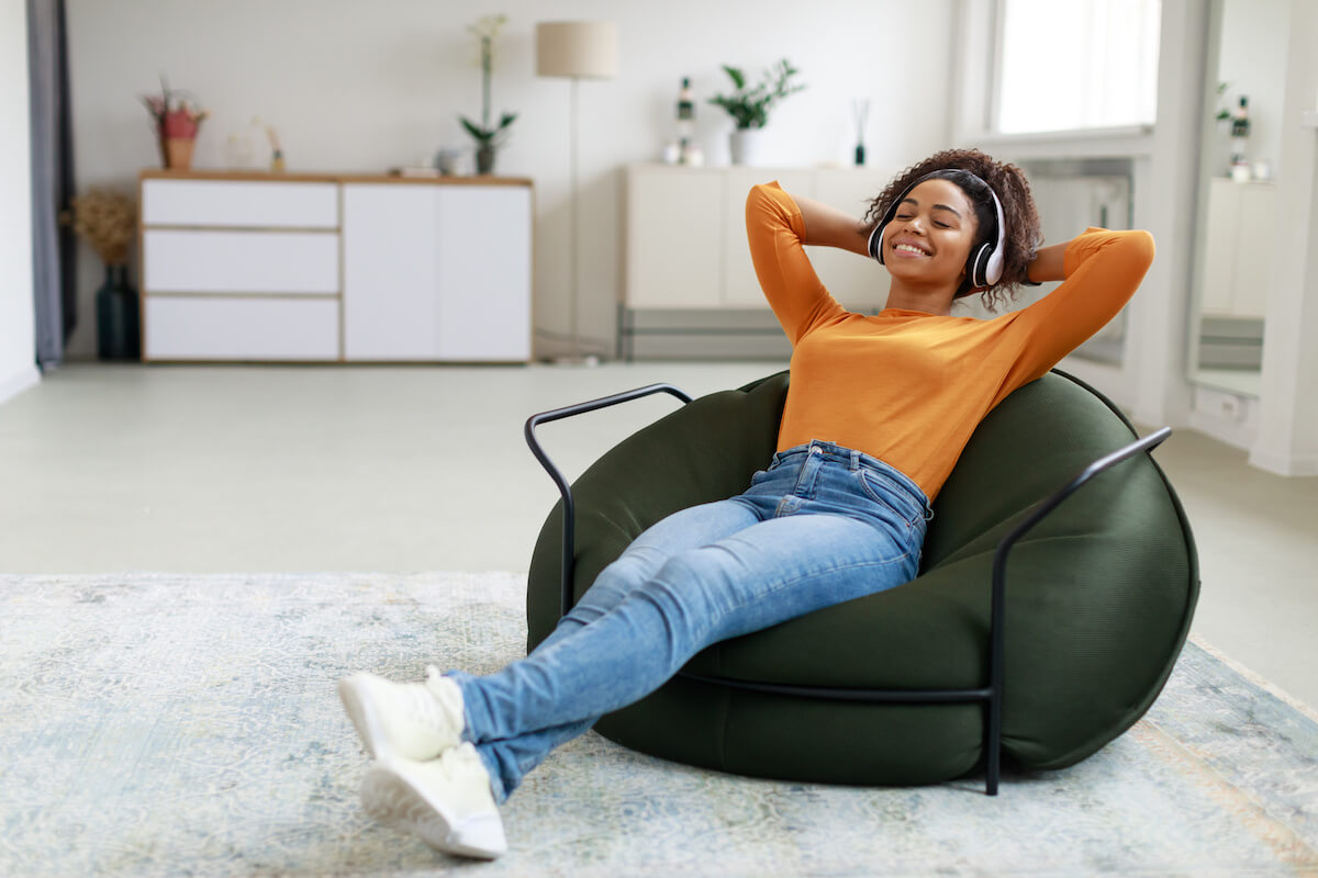 Calm black woman sitting on bean bag, listening to music, audio book, podcast, enjoying meditation for sleep and peaceful mind in wireless headphones.