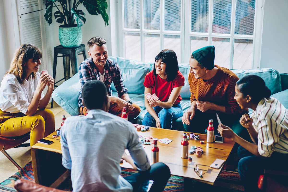 From above joyful immersed diverse hipsters playing cards with chips for poker around table and having fun in modern apartment