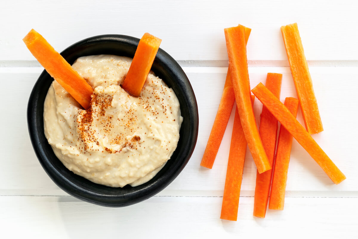 Hummus with carrot sticks, top view over white timber.