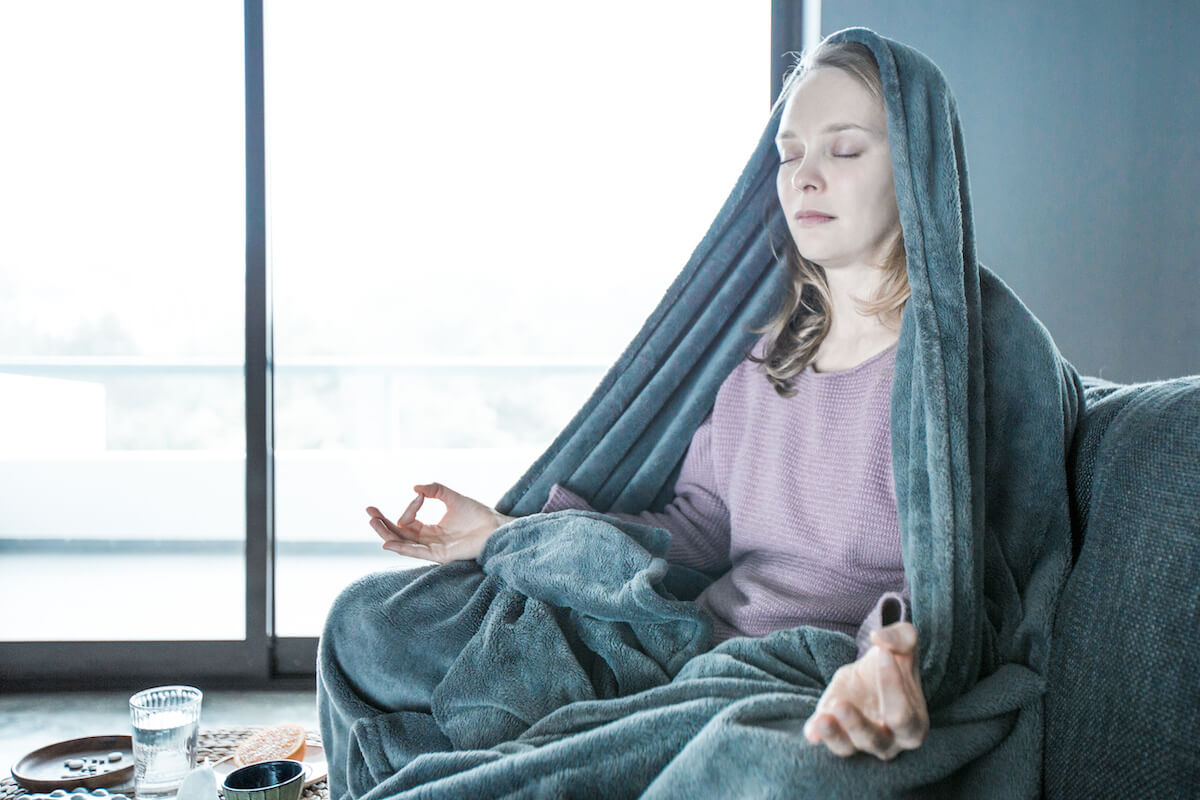 Caucasian woman in purple sweater covered with grey blanket sitting in lotus pose on grey sofa in living room, meditating