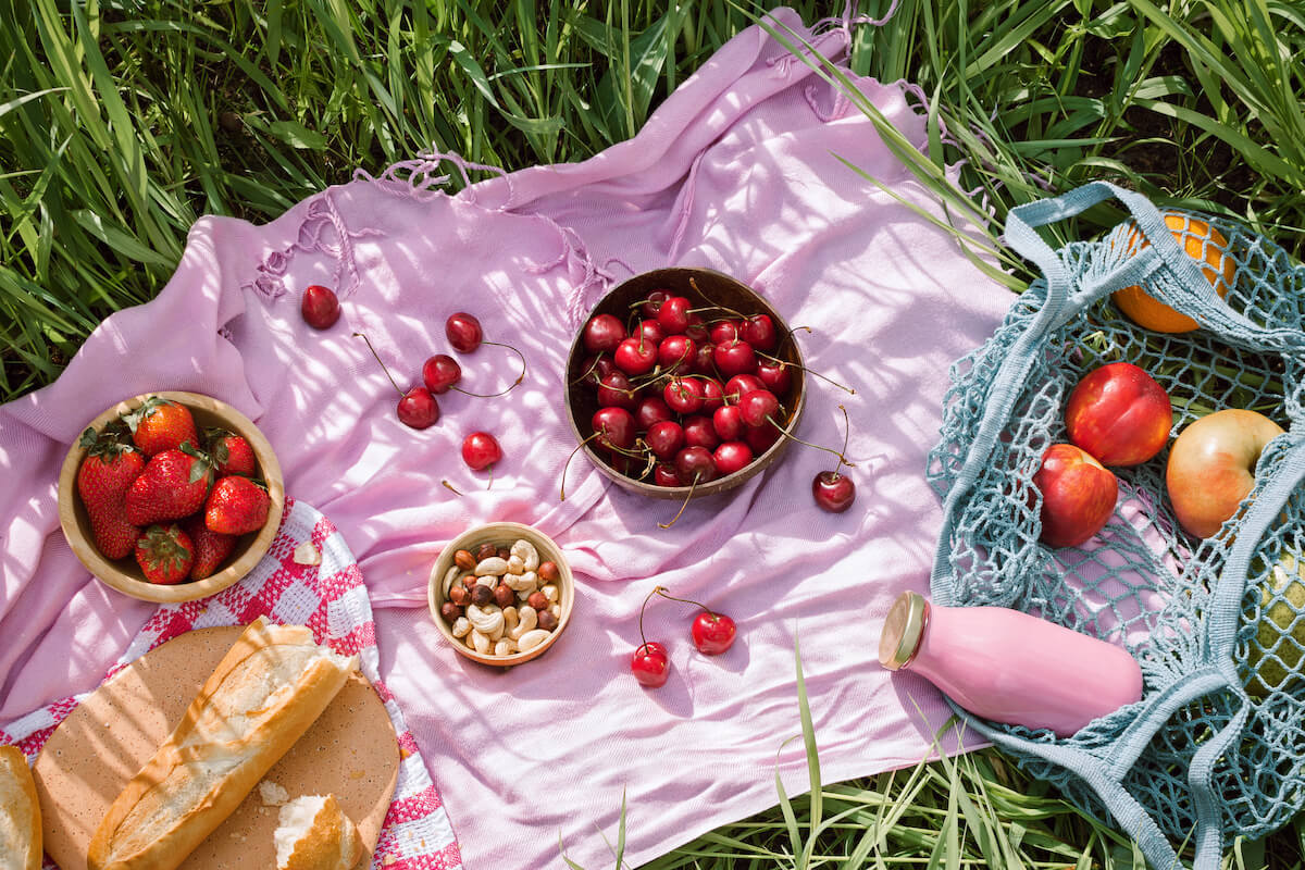 Zero waste summer picnic on the with cherries in the wooden coconut bowls, fresh bread and glass bottle of juice or smoothie on pink blanket