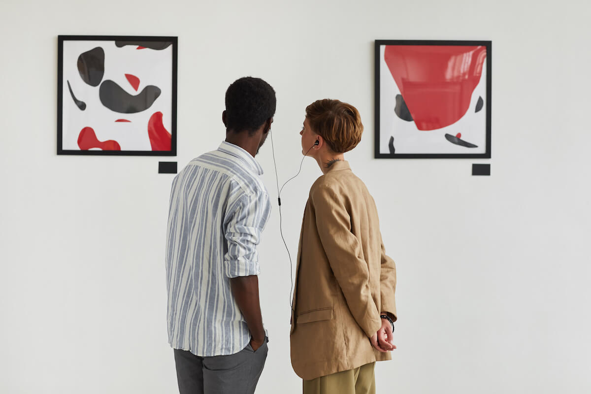 Graphic back view portrait of two young people looking at paintings and sharing audio guide while exploring modern art gallery exhibition