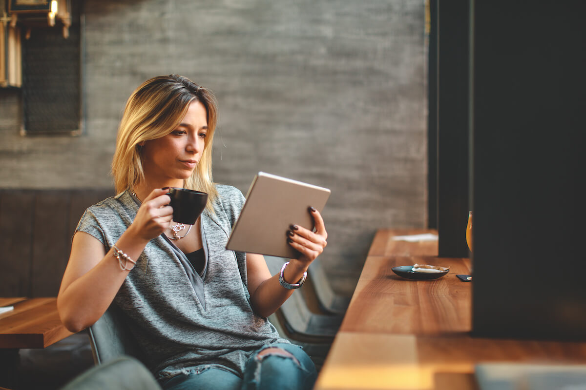 Young beautiful woman relaxing in coffee shop reading electronic book via internet on digital tablet and drink coffee