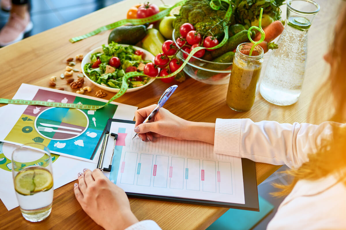 Woman dietitian in medical uniform with tape measure working on a diet plan sitting with different healthy food ingredients in the green office on background