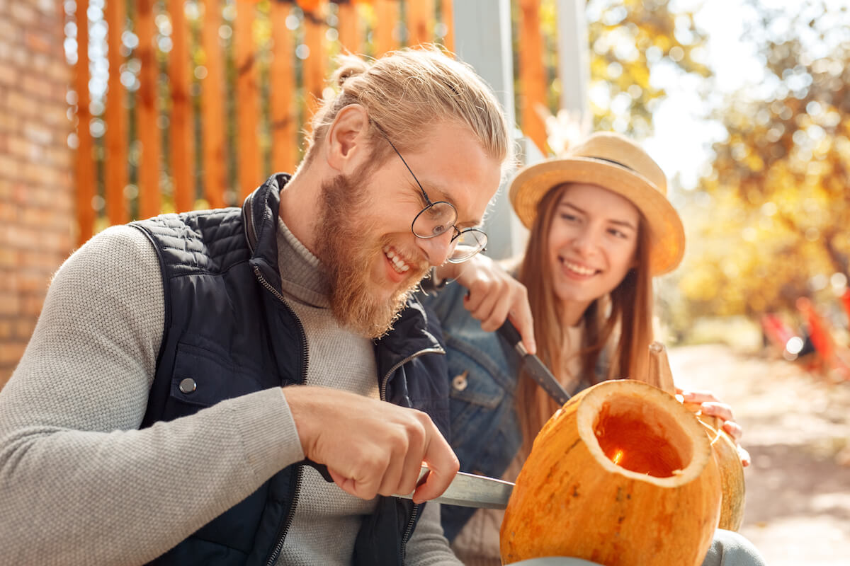 Young couple man and woman outdoors at vacation home making jack-o'-lantern preparing for halloween carving pumpkins together talking laughing cheerful close-up