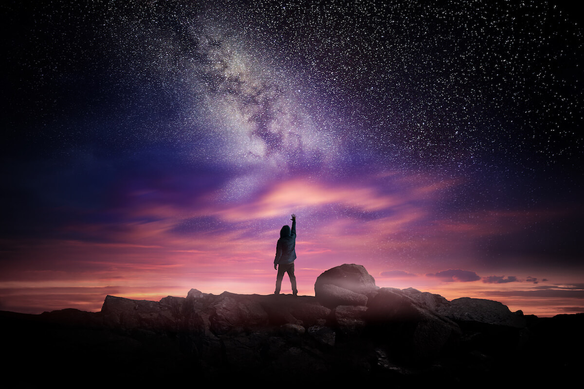 Night time long exposure landscape with a man standing in a high place reaching up in wonder to the Milky Way galaxy