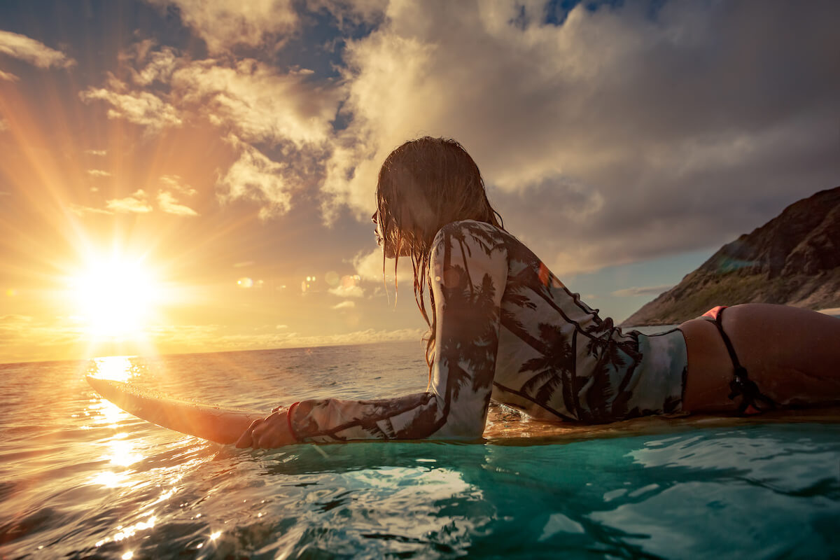 A surfer girl watching sunset on a surboard floating in blue ocean near rocky shore