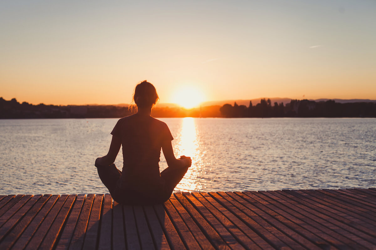 woman doing meditation and breathing exercises on wooden pier near lake, silhouette at sunset