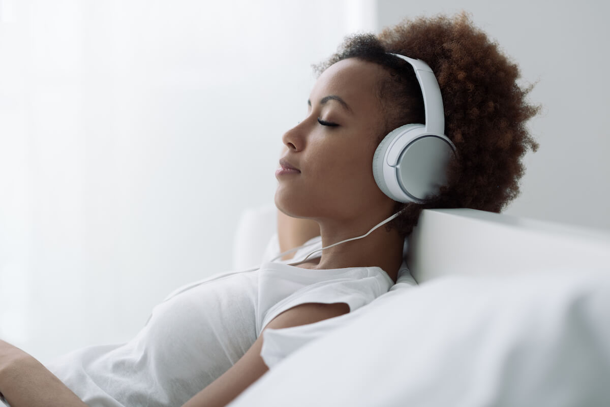 Young beautiful african american woman relaxing and listening to music using headphones, she is lying in bed