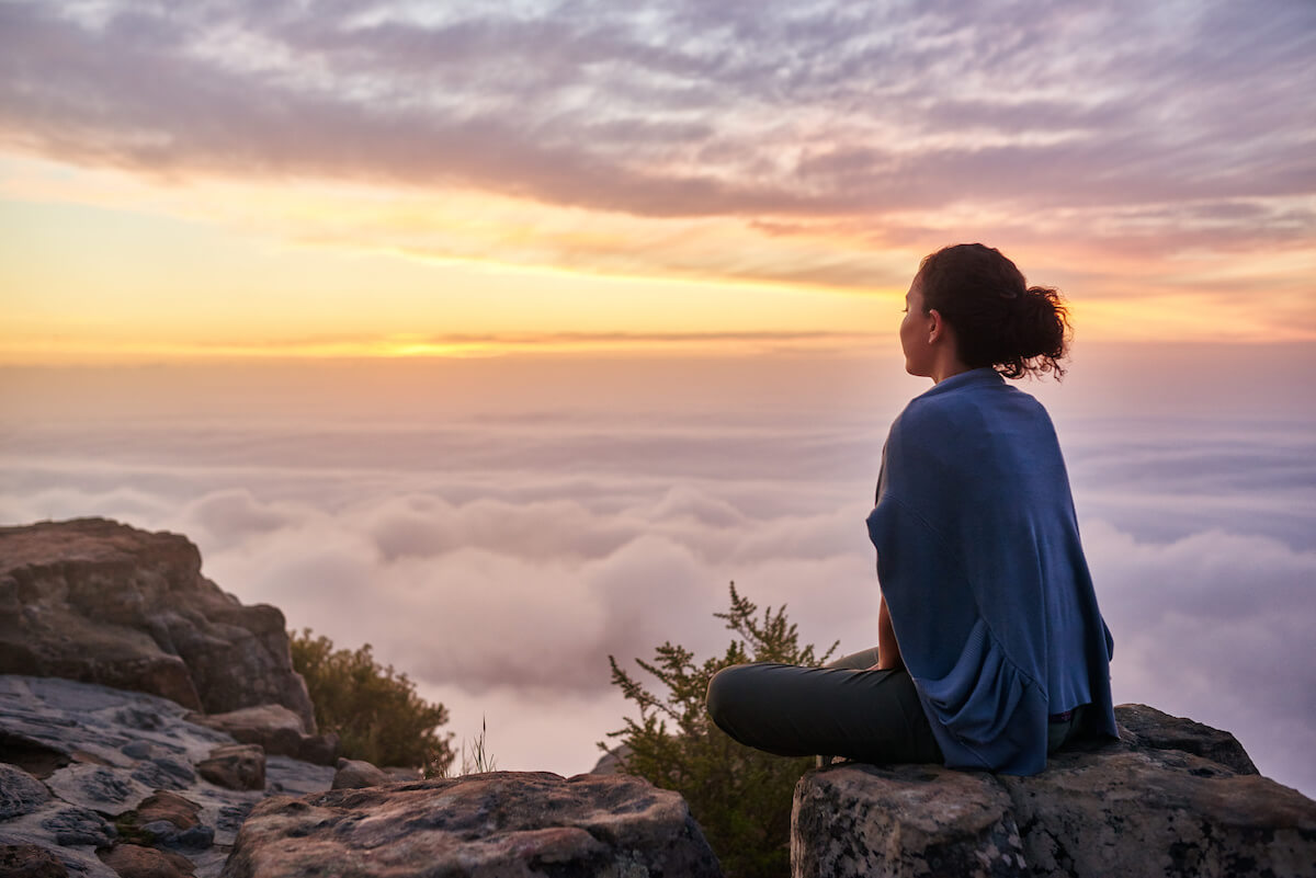 Rearview of a young woman sitting on a mountain top peacefully gazing at low-lying morning clouds and the pastel colours of a tranquil sunrise