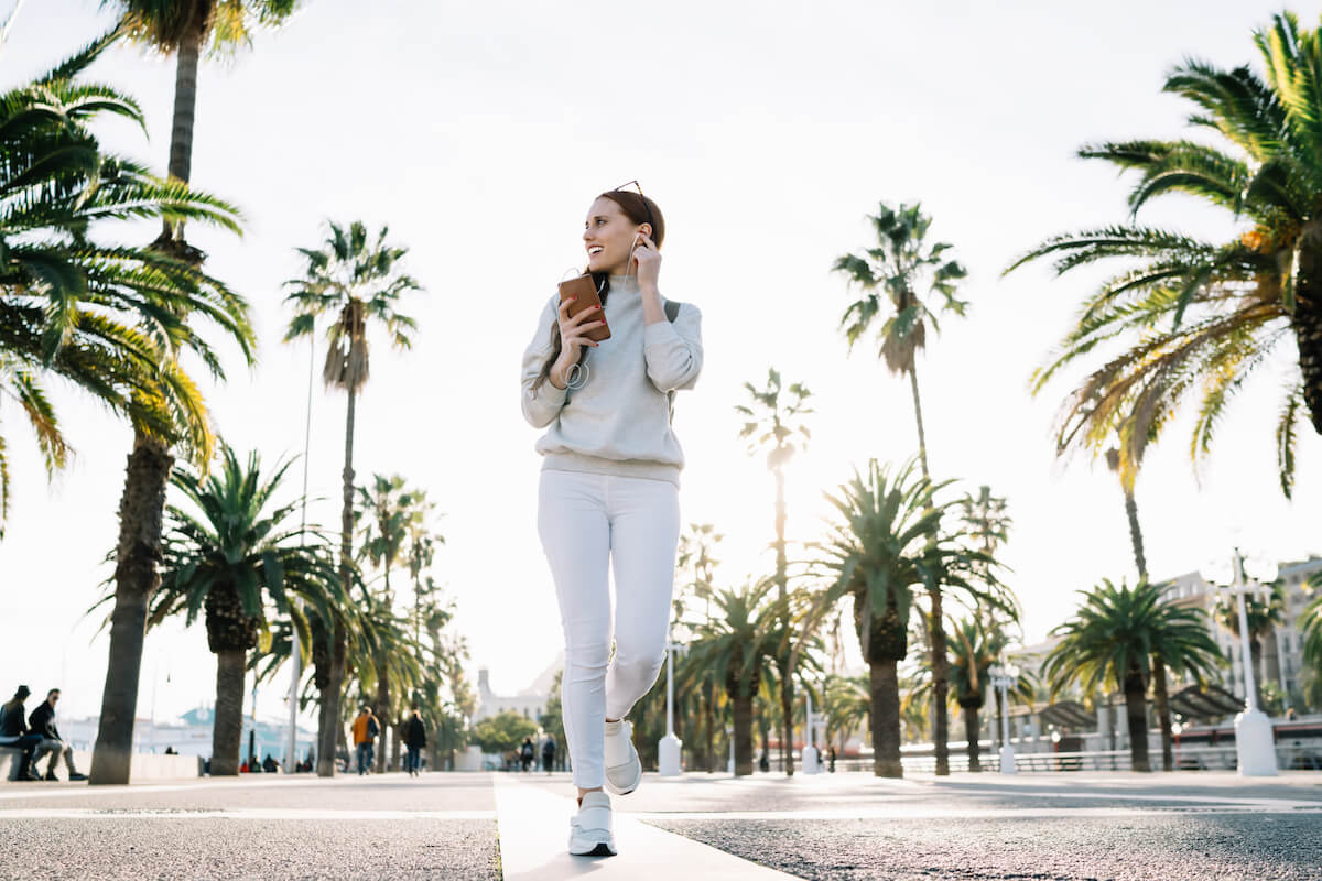 From below pleased pensive attractive adult female in white clothes touching on headphone and looking away while walking and talking on phone in park during sunny day on blurred tropical background