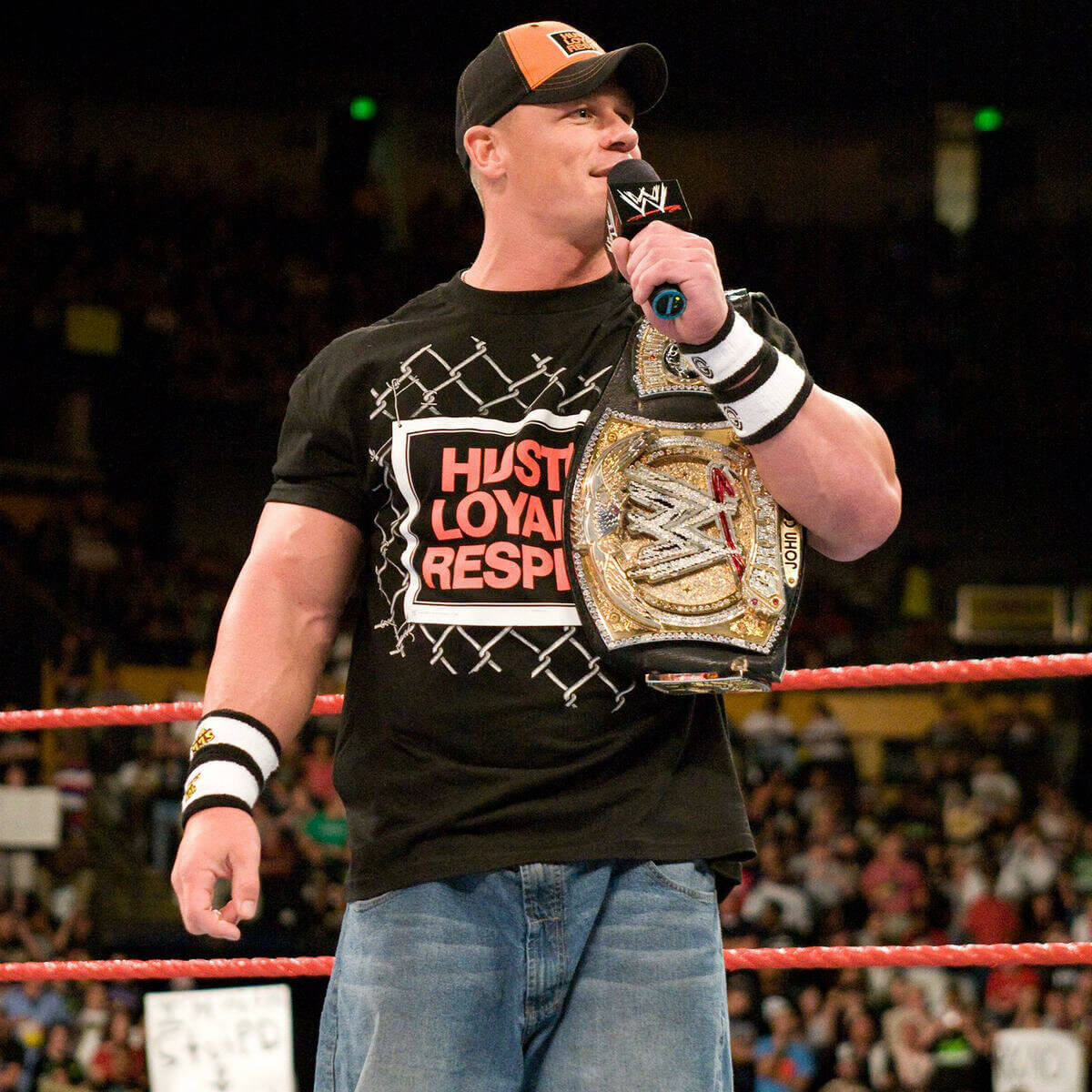 John Cena talking on mic in a boxing ring holding a champion belt. 